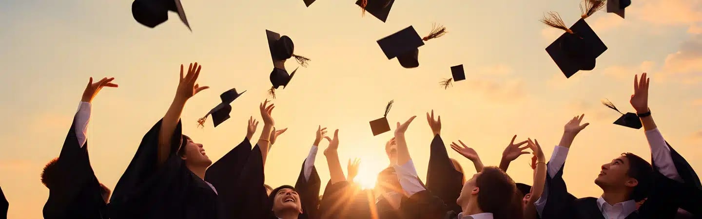 Graduates in caps and gowns joyfully toss their caps in the air during a sunset celebration.