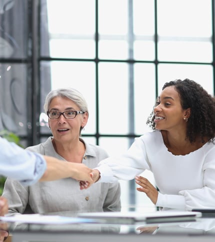 Two women sit at a table in a bright, modern office—one, a Social Service Assistant with gray hair and glasses looks surprised, while the other smiles and leans forward to shake hands with someone out of frame.
