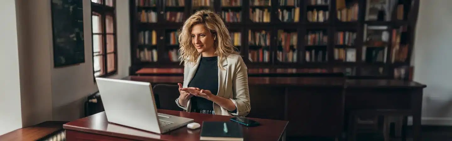 A woman in a library gestures while video conferencing on a laptop, with bookshelves in the background.