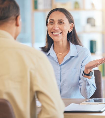 A woman in a light blue shirt smiles and gestures while sitting at a table with a man, suggesting a friendly conversation or interview in a bright, modern office where she works as a Legal Support Worker.