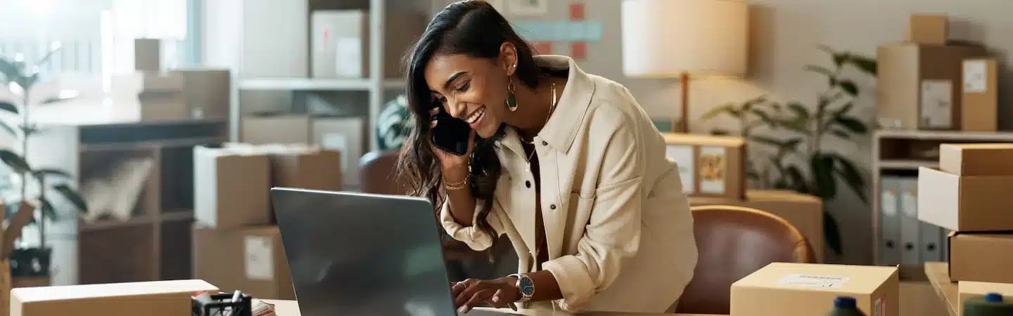 A woman smiles while talking on the phone and using a laptop in a room filled with boxes.