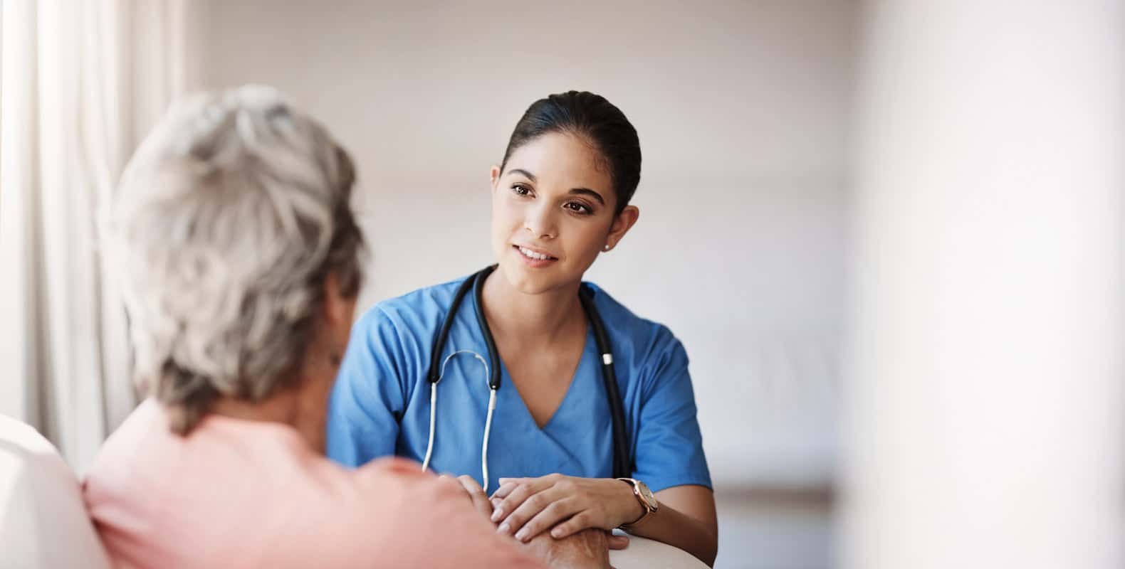 A healthcare worker in blue scrubs and a stethoscope smiles while talking to an older person. The setting appears calm and supportive, suggesting a medical consultation or caregiving environment.