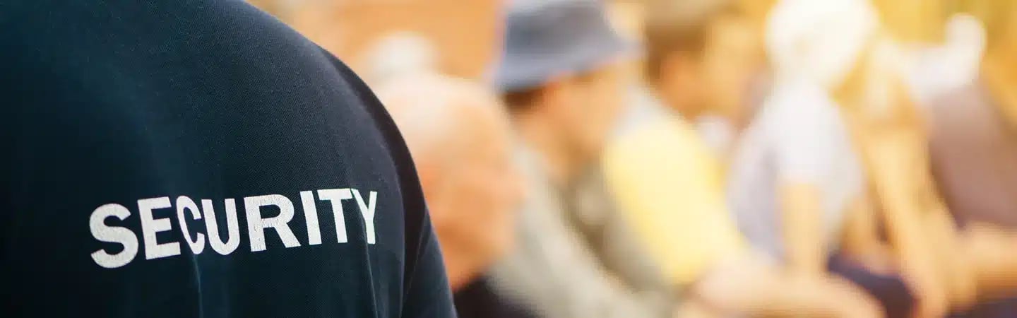 Close-up of a person wearing a shirt labeled "Security" with a blurred background of seated people.