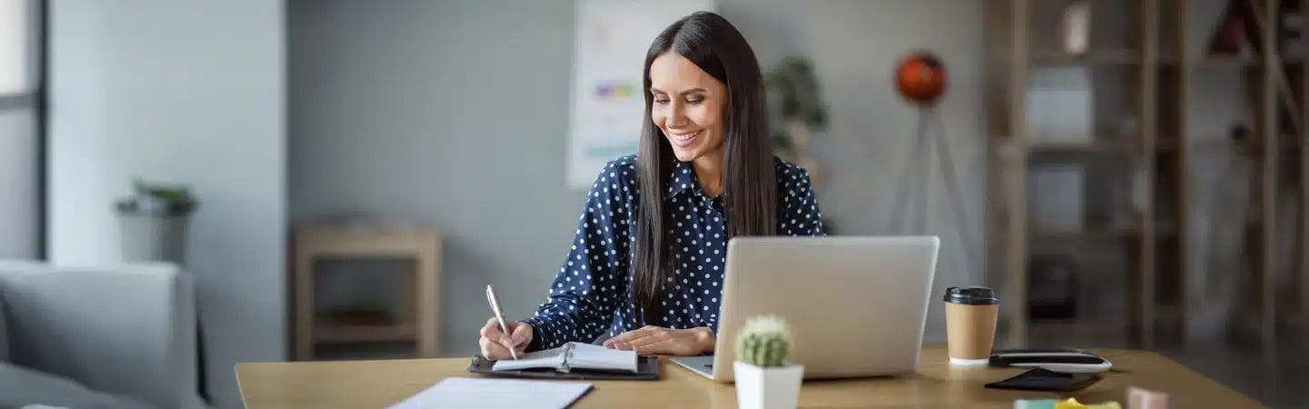A woman smiles while working at a desk with a laptop, notebook, and coffee cup in a bright, modern office.