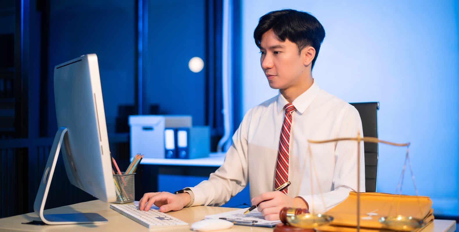 A young man in a white shirt and striped tie works at a computer in an office at night, with a notepad, scales of justice, and stationery on the desk.