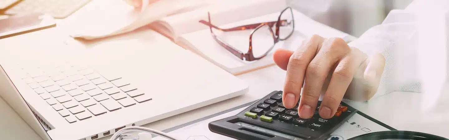 Person using a calculator at a desk with a laptop, documents, and eyeglasses.