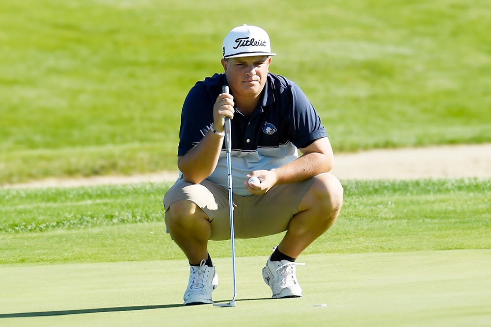 Golfer in a cap and shorts crouching on a green, reading the line for a putt in a sunny outdoor golf course.