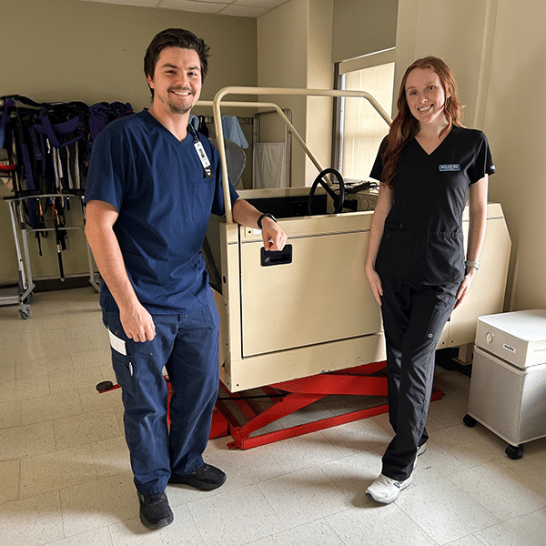 Two healthcare workers in scrubs stand beside a medical device in a clinical setting, smiling at the camera.