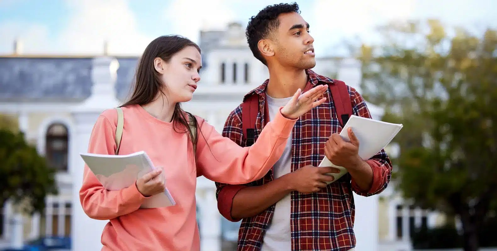 Two students holding notebooks, looking around with a building in the background on a sunny day.