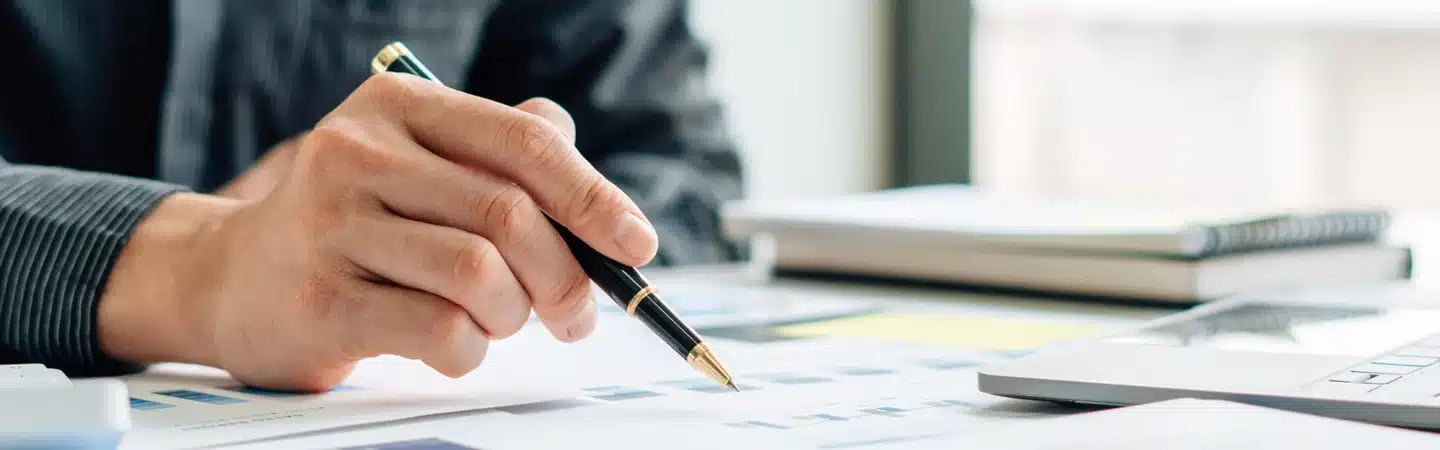 A person holding a pen reviews documents on a desk with a laptop and notebooks in the background.