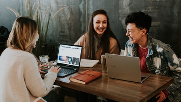 Three people laughing and working on laptops at a table, with notebooks and a plant in the background.