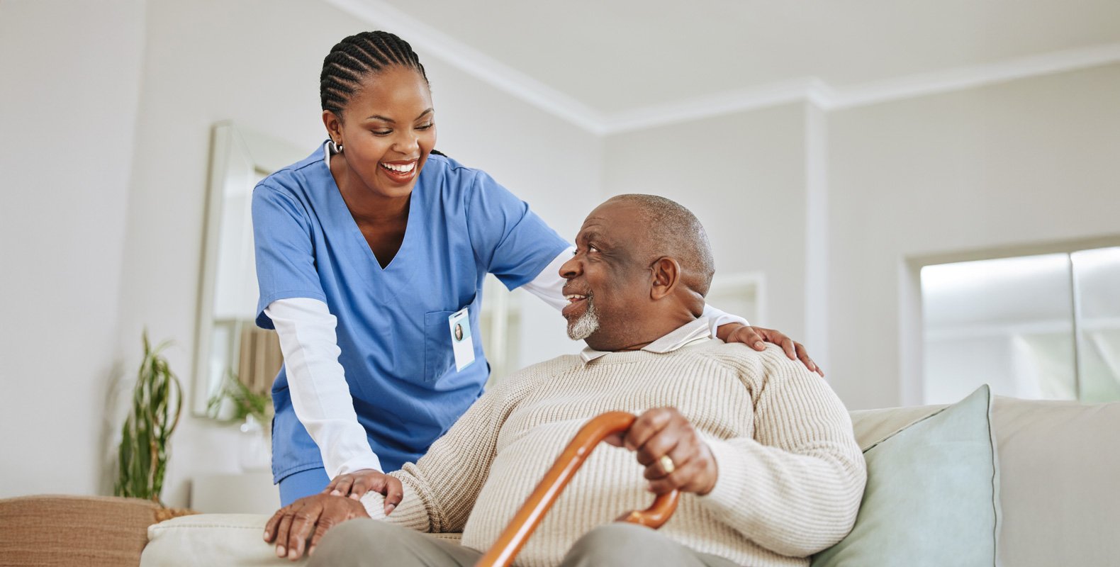 A smiling healthcare worker in blue scrubs stands beside an elderly man with a cane, who is sitting on a couch. They appear to be enjoying a friendly conversation in a bright, comfortable living room.