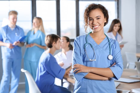 A smiling healthcare worker in blue scrubs with a stethoscope stands in the foreground, while a group of medical professionals converse and work together in the background of a bright room.