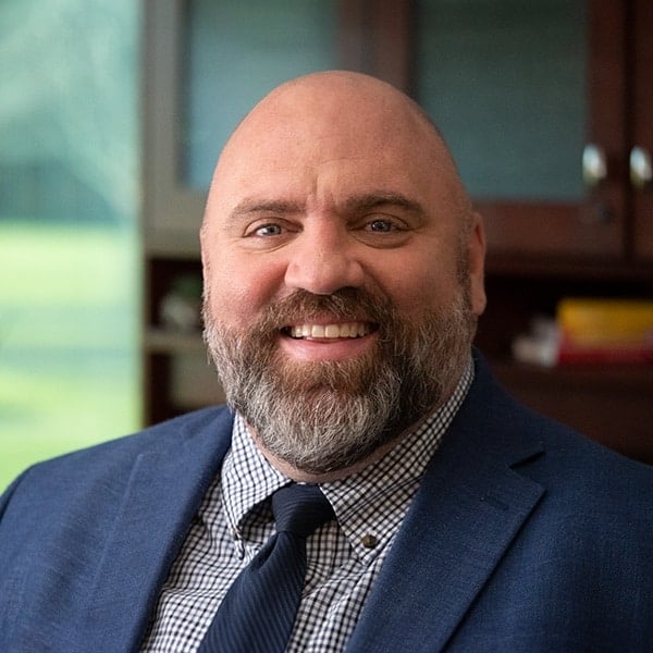 Smiling man with a bald head and a beard, wearing a suit and tie, sitting in an office.