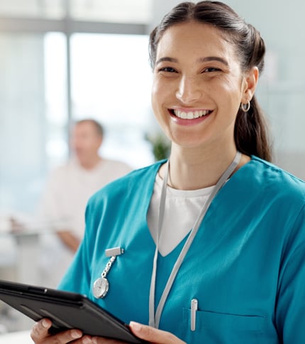 A smiling Medical Assistant wearing teal scrubs and a lanyard holds a tablet in a bright medical setting, with a blurred patient in the background.