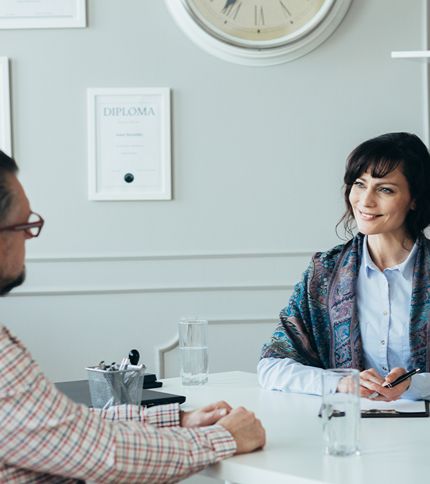 A woman smiles while talking to a Human Resources Specialist across a desk in an office setting. There are diplomas on the wall, a clock, and glasses of water on the desk.