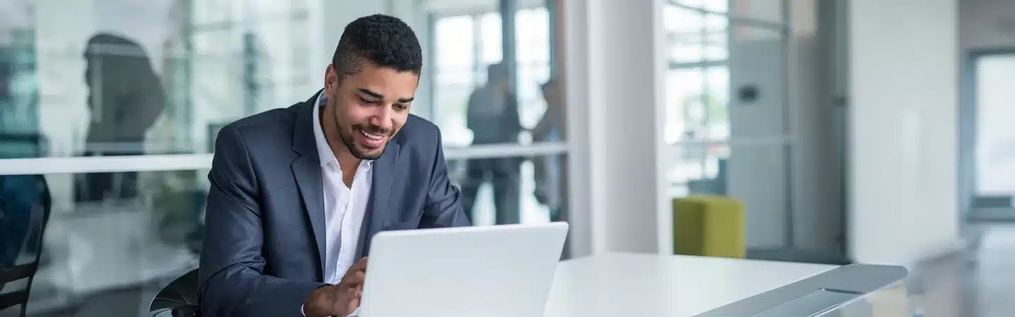 Man in a suit smiles while working on a laptop at an office desk with glass walls in the background.