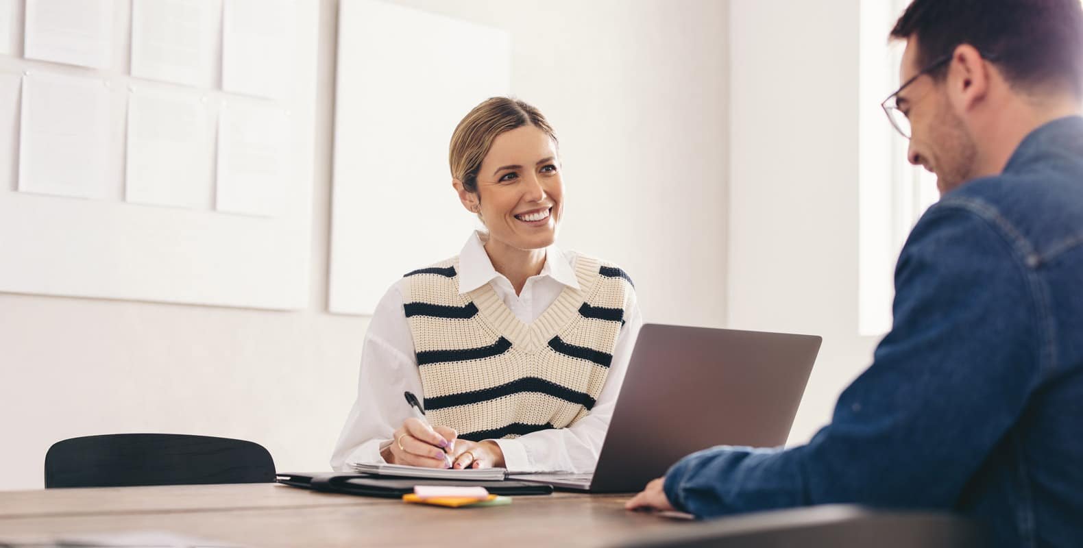 A woman is smiling and sitting at a table with a laptop and notebook, looking at a man seated across from her. They appear to be having a meeting in a bright room with white walls and paper documents on a board.