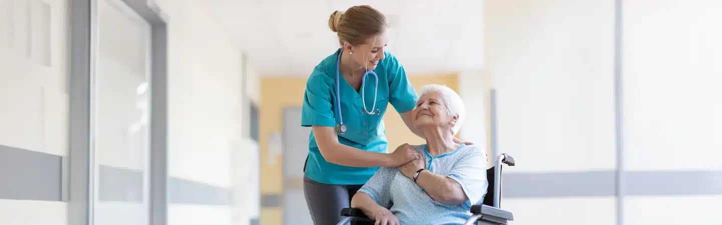 Nurse smiling at elderly woman in a wheelchair in a hospital hallway.