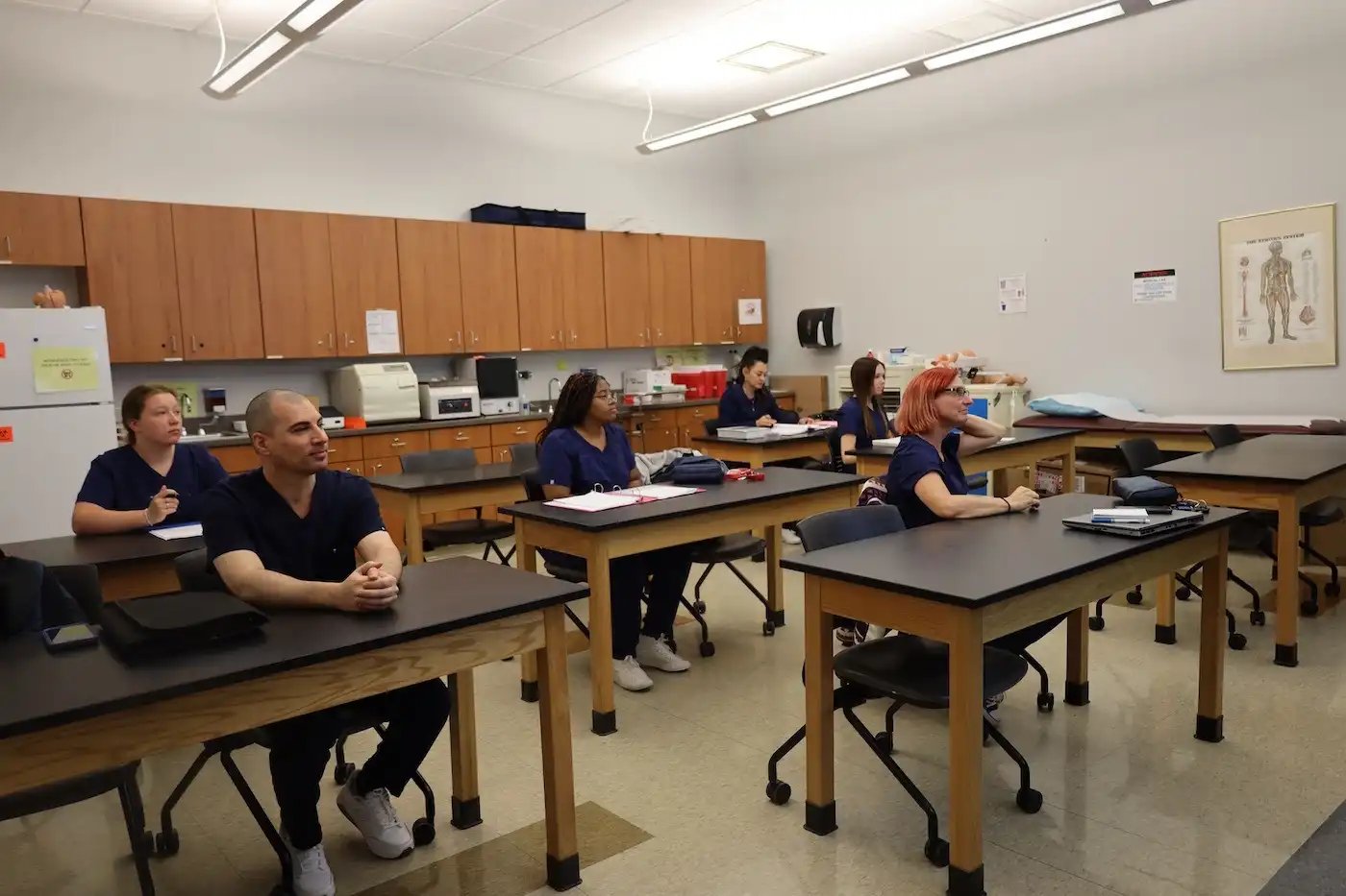 A classroom with six students in matching blue scrubs sitting at desks, attentively listening to a lecture.