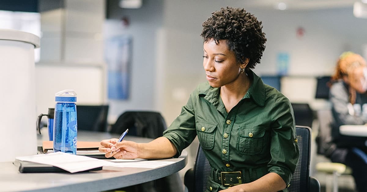 A woman in a green shirt sits at a desk with a pen, writing in a notebook, with a blue water bottle beside her.
