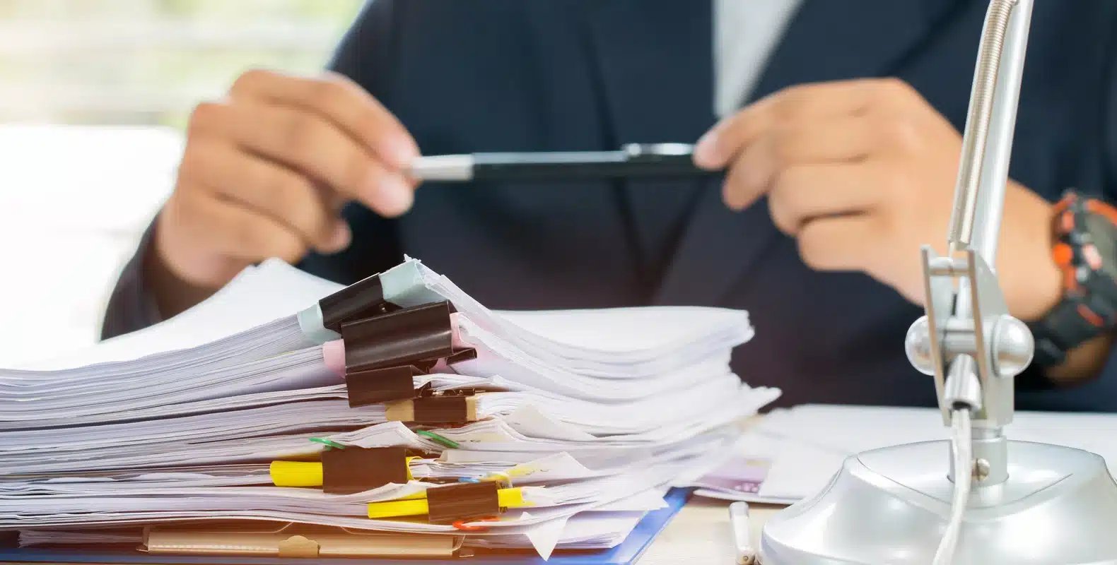 Person holding a pen, sitting at a desk with a large stack of documents clipped together and a desk lamp nearby.
