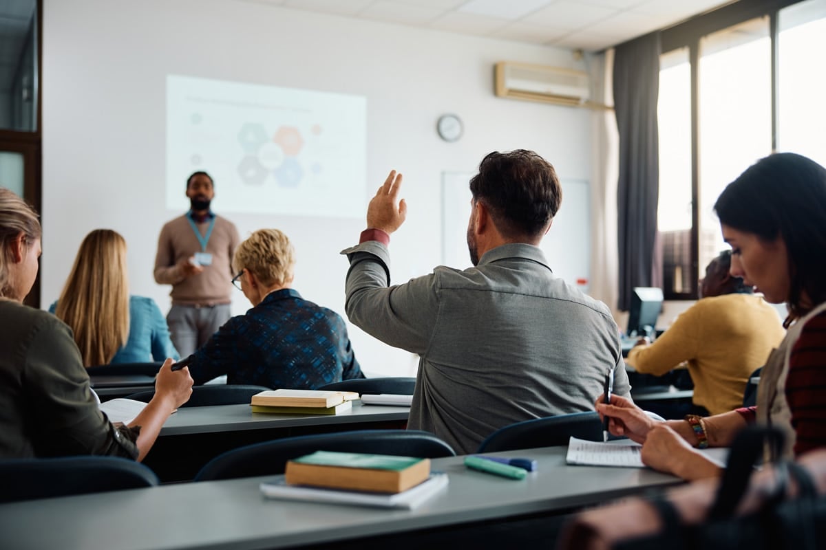 A man raises his hand during a classroom lecture with a projector screen in the background and seated students.
