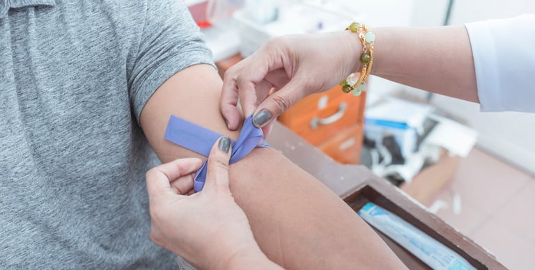 A person wearing a gray shirt is having a tourniquet tied on their upper arm by someone preparing for a medical procedure, likely a blood draw, in a clinical setting.