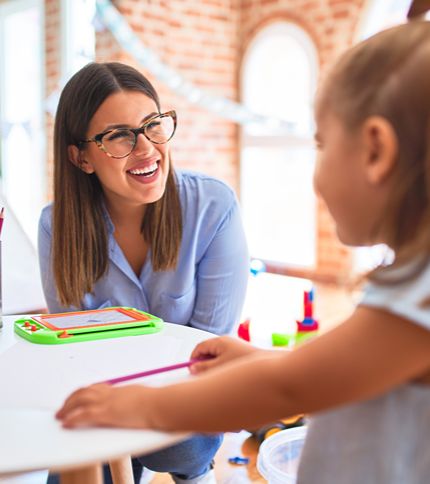 A Daycare Center Administrator wearing glasses smiles while sitting at a table with a young child holding a colored pencil. Art supplies and toys are spread out in the bright, cheerful room.