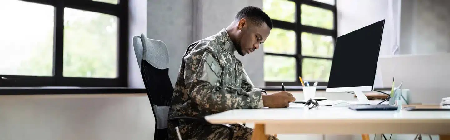 A person in military uniform sits at a desk, writing in a notebook beside a computer in a well-lit office.