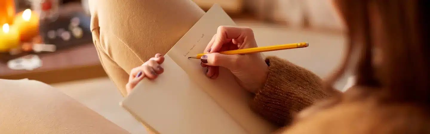 Person writing in a journal with a pencil, cozy warm lighting and candle-lit background.