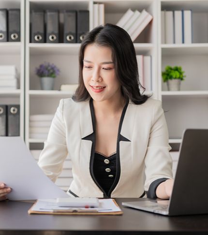An Accounts Payable Clerk in a white blazer sits at a desk, holding a paper in one hand and working on a laptop with the other. Behind her are shelves with books, folders, and small potted plants.