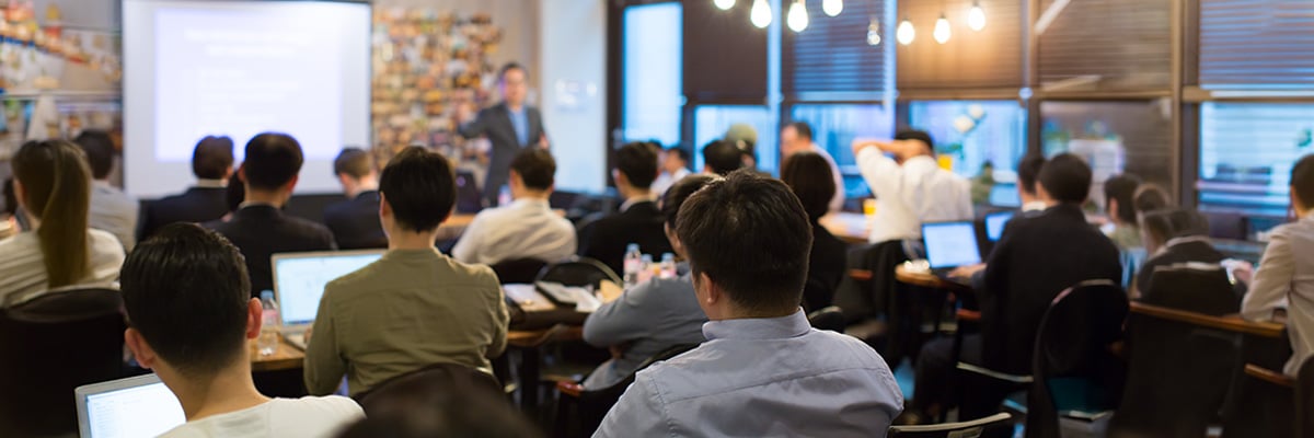 A group of people are seated in a classroom-style setting, engaged in professional development as they listen to a presenter at the front. Most attendees face forward, some using laptops, with a presentation projected on a screen.