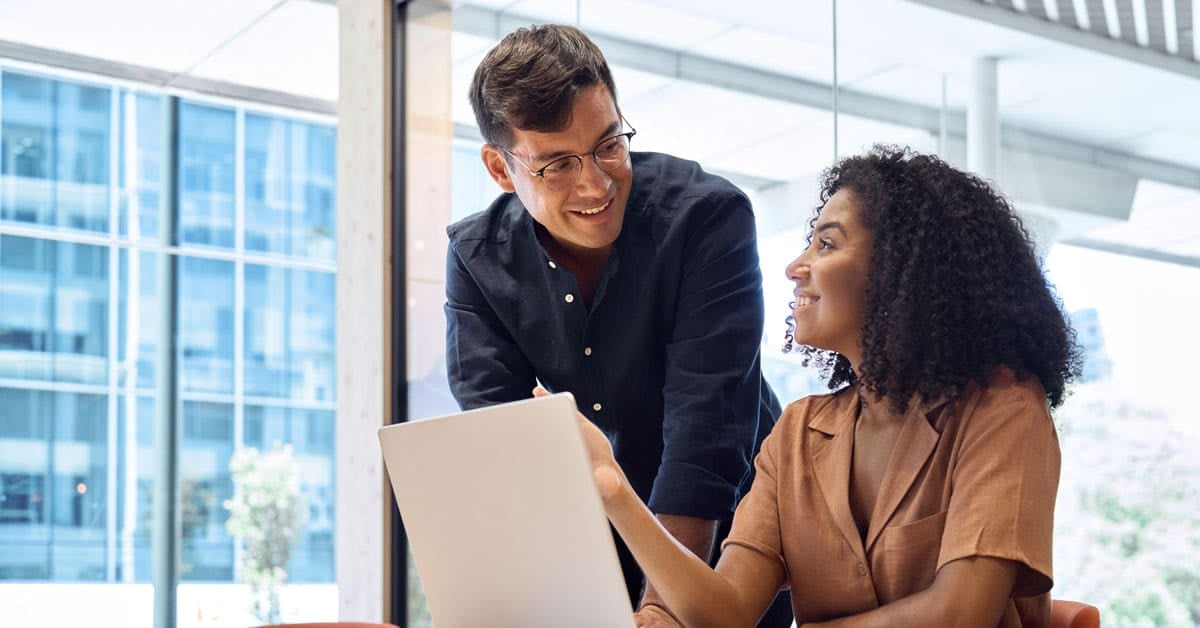 Two colleagues in Organizational Management share a friendly conversation in a modern office. One sits at a desk using a laptop while the other stands beside her, both smiling in front of large windows.