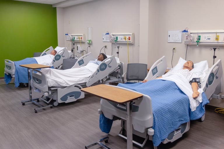 Three hospital beds with medical mannequins covered by blue blankets in a clinical training room in Orchard Park, NY, with medical equipment and supplies on the walls behind the beds.