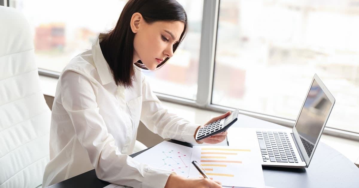A woman at a desk using a calculator and writing on documents, with a laptop open beside her.
