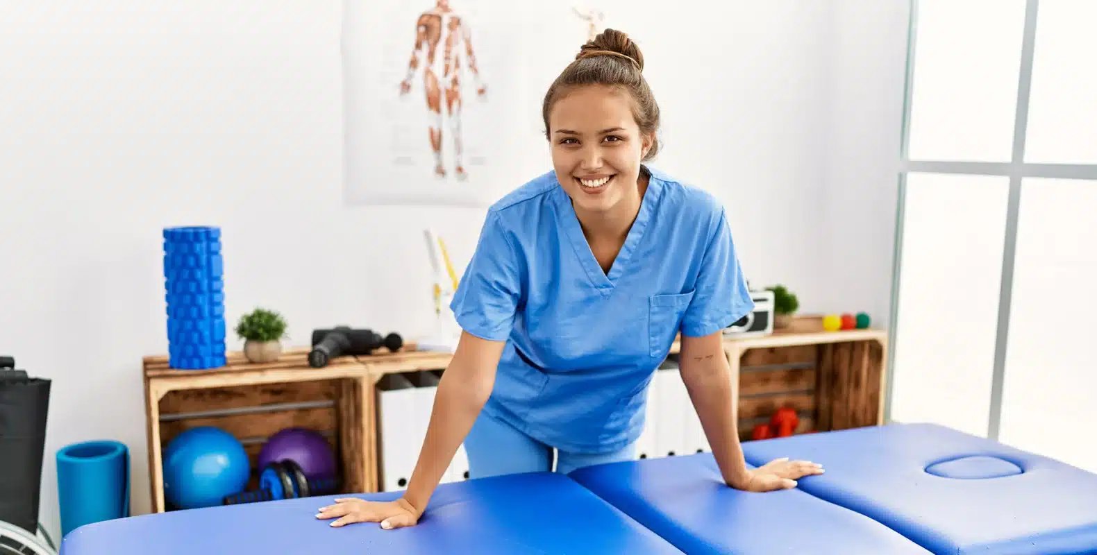 A smiling person in blue scrubs stands beside a massage table in a therapy room filled with exercise equipment.
