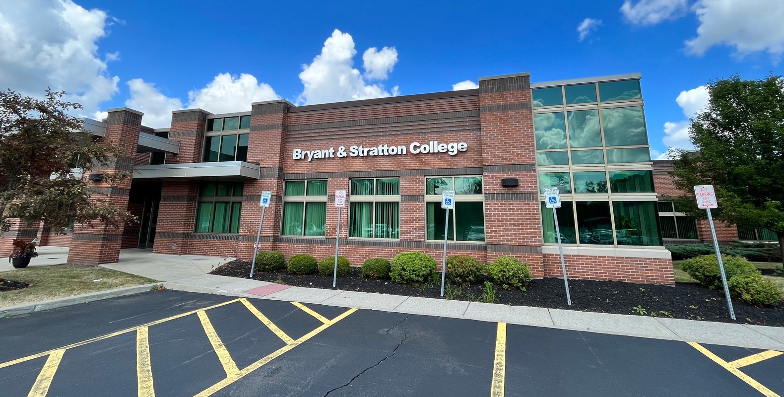 A modern brick building with large windows and a sign reading "Bryant & Stratton College" on the front. There are accessible parking spaces and green shrubs in front, under a blue sky with clouds.