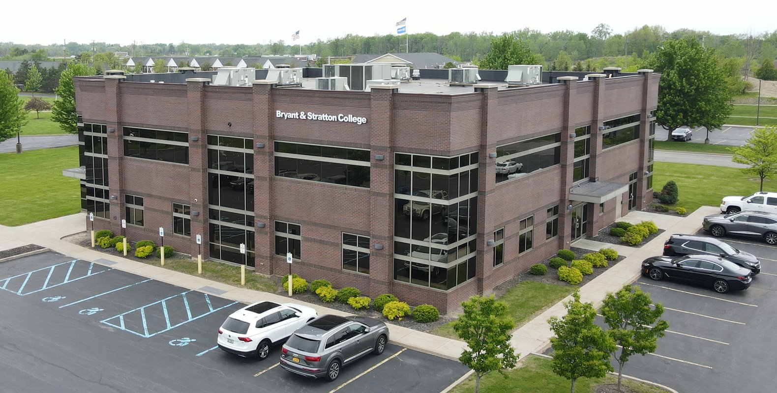 A two-story brick building with large windows shows a sign reading "Bryant & Stratton College" in Orchard Park, NY. Several cars are parked outside, including accessible spaces. Green trees and grass surround the building.