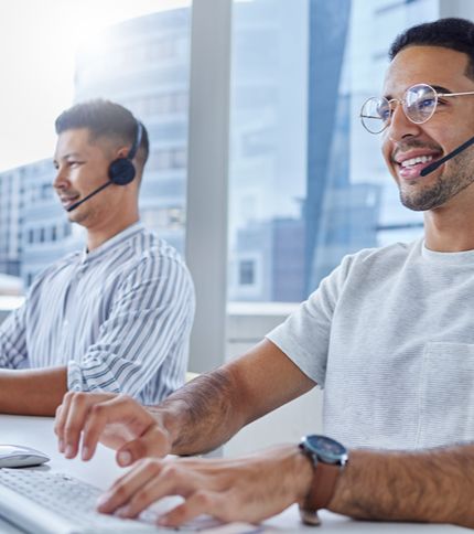 Two men wearing headsets and casual shirts sit at desks, smiling as they handle customer service responsibilities in a bright, modern office with large windows and city buildings visible outside.