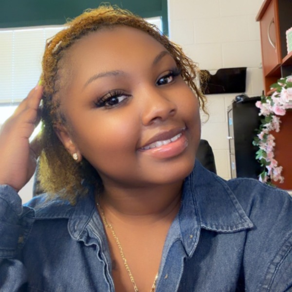 A smiling woman with shoulder-length blonde dreadlocks wearing a blue denim shirt.