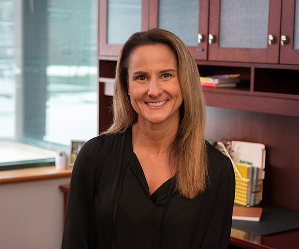A woman with straight, light brown hair and a black blouse smiles while sitting in an office with wooden shelves, books, and a window in the background at Bryant & Stratton College.