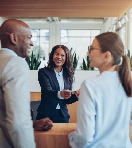 A receptionist in a business suit smiles while handing a key card to two people standing at a reception desk in a modern office or hotel lobby with green plants in the background.