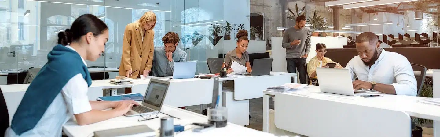 A diverse group of professionals working on laptops and documents in a modern, open-plan office setting.