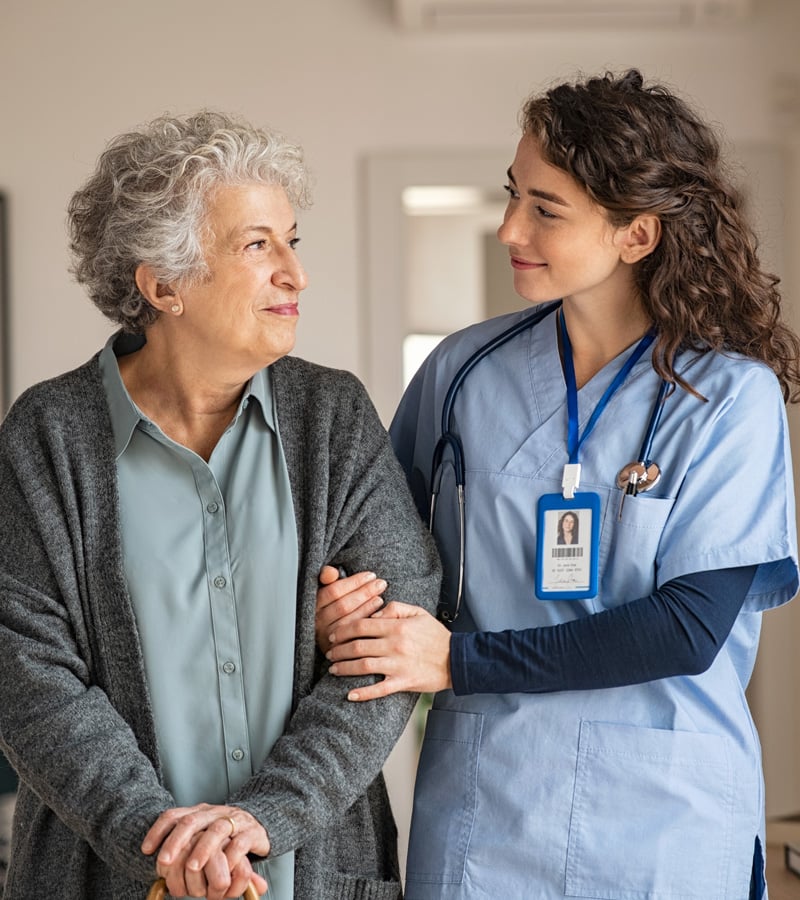 An elderly woman with gray hair smiles while holding a cane, standing beside a nurse in blue scrubs who supports her arm and smiles warmly at her.