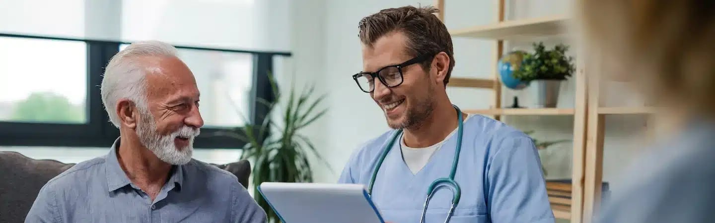 A smiling older man sits with a doctor in scrubs holding a tablet, both engaged in a friendly conversation.