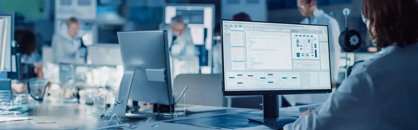 Person in a lab coat working at a computer in a laboratory with screens displaying complex data and analysis.