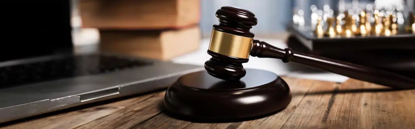 A judge's gavel on a wooden desk with a laptop, books, and a chess set in the background.