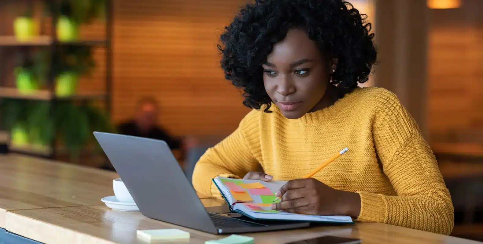 Person in a yellow sweater working at a laptop and writing in a notebook, seated at a wooden table in a cozy setting.