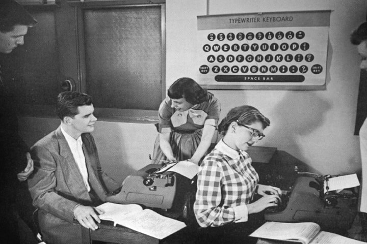 Three people observe a woman typing on a typewriter in a classroom with a large keyboard chart on the wall.